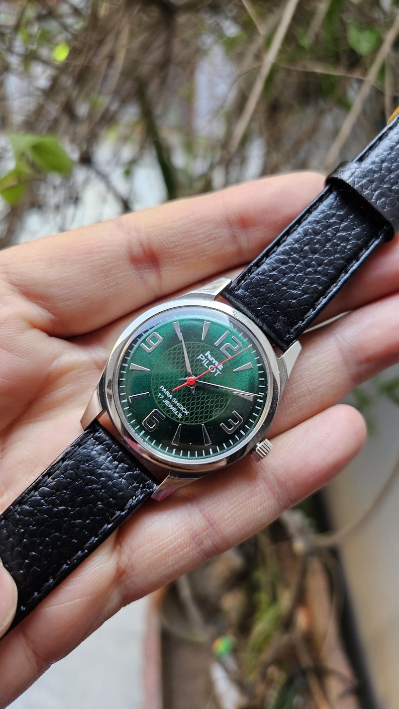 May include: A silver wristwatch with a green face and black leather band. The watch face has white numbers and a red second hand. The text on the watch face reads "Time Pilot Para Shock 17 Jewels".