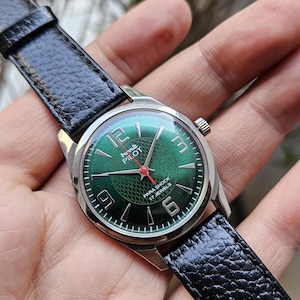 May include: A green-faced wristwatch with a black leather band. The watch face has white numerals and a red second hand. The watch face also has the text "HMT PILOT PARA SHOCK 17 JEWELS".