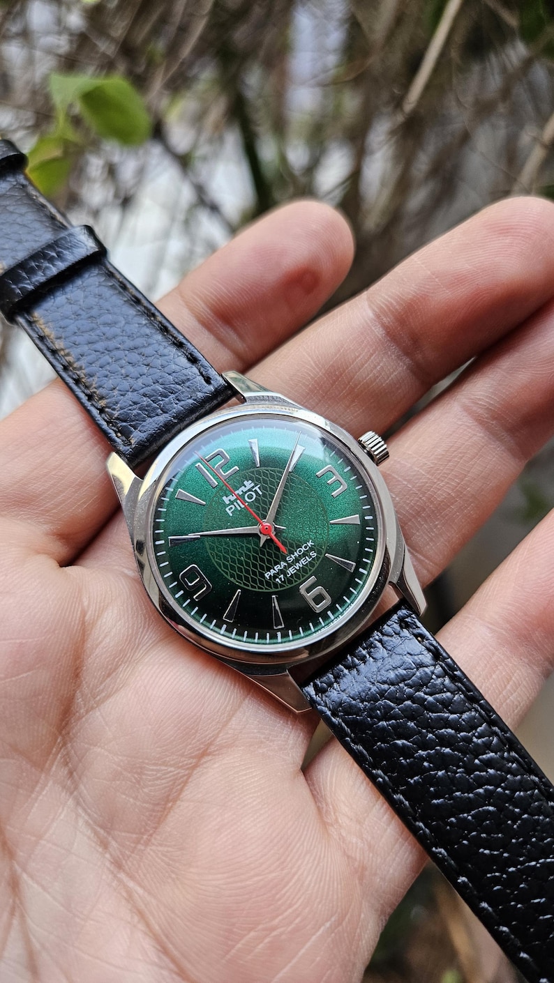 May include: A green-faced wristwatch with a black leather band. The watch face has white numerals and a red second hand. The watch face also has the text "HMT PILOT PARA SHOCK 17 JEWELS".