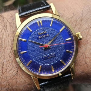 Könnte beinhalten: Eine goldfarbene Armbanduhr mit blauem Zifferblatt und schwarzem Lederarmband. Das Zifferblatt der Uhr hat ein Gittermuster und den Text "HMT Sona Para Shock 17 Jewels".