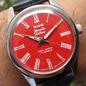 May include: A red-faced wristwatch with silver hands and Roman numeral markings. The watch face reads "hmt Janata Deluxe Para Shock 17 Jewels Made in India 0231".