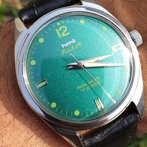 Op de afbeelding: Een polshorloge met een groene wijzerplaat en een zwart leren bandje. De wijzerplaat van het horloge heeft een zilveren rand en witte wijzers. Op de wijzerplaat van het horloge staan de woorden "humt Pilot" en "PARA SHOCK 17 JEWELS" gedrukt.