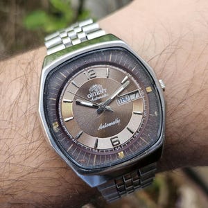 May include: A silver wristwatch with a brown face and a silver metal band. The watch face has a date window that says "THU 7" and the words "ORIENT Automatic" printed on it.