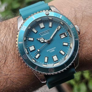 Può includere: Un orologio automatico Citizen con quadrante e lunetta turchese, indici delle ore rettangolari bianchi e visualizzazione della data. L'orologio ha un cinturino in gomma turchese e una cassa color argento. Il testo "WATER RESISTANT 150M" è visibile.