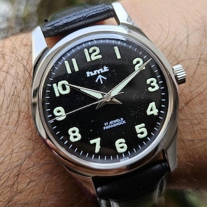 May include: A stainless steel wristwatch with a black face and a black leather band. The watch face features white numerals, luminous hands, and the text "hmt" and "17 JEWELS PARASHOCK". The watch is approximately 1.5 inches in diameter.