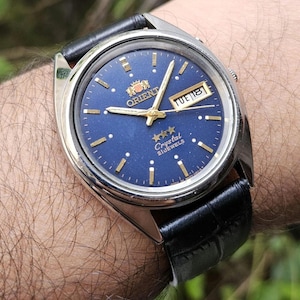 May include: A close-up of an Orient watch with a blue face and gold accents. The watch has a silver case and a black leather band. The watch face displays the day and date. The watch is a vintage style.