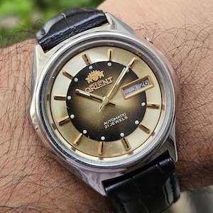May include: An Orient automatic wristwatch with a silver-tone stainless steel case and a black leather strap. The dial features a gold-to-brown gradient, gold-tone hour markers, luminous dots, and a day-date window displaying the number 26.