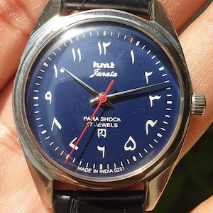 May include: A blue-faced wristwatch with Arabic numerals, a red second hand, and the words "HMT Janata Para Shock 17 Jewels Made in India 0231".