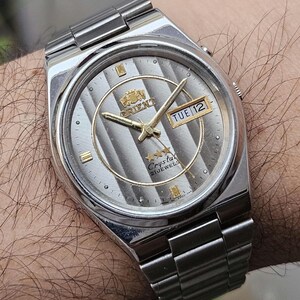 May include: Silver-toned Orient Crystal 21 Jewels wristwatch with a textured silver dial and gold-colored hands and accents. The watch displays the day and date. The watchband is made of silver-toned metal.