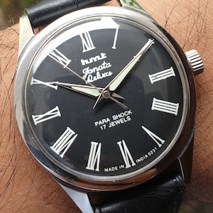 Könnte beinhalten: Eine schwarze Armbanduhr mit einem silbernen Gehäuse und einem schwarzen Lederarmband. Das Zifferblatt der Uhr hat römische Ziffern als Stundenmarkierungen, einen weißen Sekundenzeiger und den Text "hmt Janata Deluxe Para Shock 17 Jewels Made in India 0231".