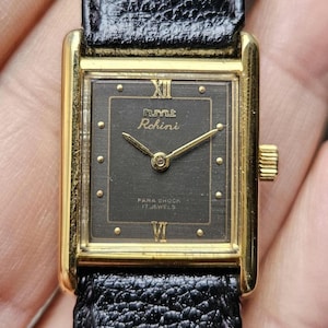 Könnte beinhalten: Eine goldfarbene Armbanduhr mit schwarzem Zifferblatt und schwarzem Lederarmband. Das Zifferblatt der Uhr hat goldfarbene Zeiger und römische Ziffern als Stundenmarkierungen. Das Zifferblatt der Uhr trägt außerdem den Text "Rhm & Rohini" und "Para Shock 17 Jewels".
