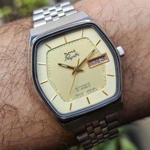 Puede incluir: Un reloj de pulsera automático HMT Pragathi vintage con caja y correa plateadas. La esfera del reloj es de color oro claro con manecillas e indicadores dorados. El día se muestra como "DOM 3". El reloj indica "AUTOMATIC 21 JEWELS INDIA 83 6090020".