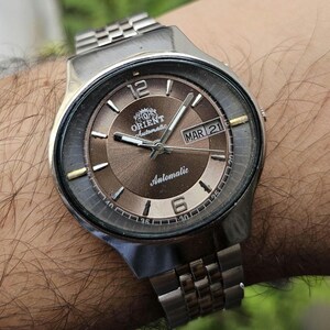 May include: An Orient automatic wristwatch with a metallic brown sunburst dial, silver-tone hands, and hour markers. The watch features a day-date window at the three o'clock position, the text Orient Automatic, and a stainless steel link bracelet.