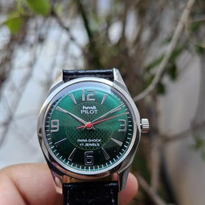 May include: A green-faced wristwatch with a silver case and black leather band. The watch face has white numerals and a red second hand. The watch face also has the text "hmt PILOT PARA SHOCK 17 JEWELS".