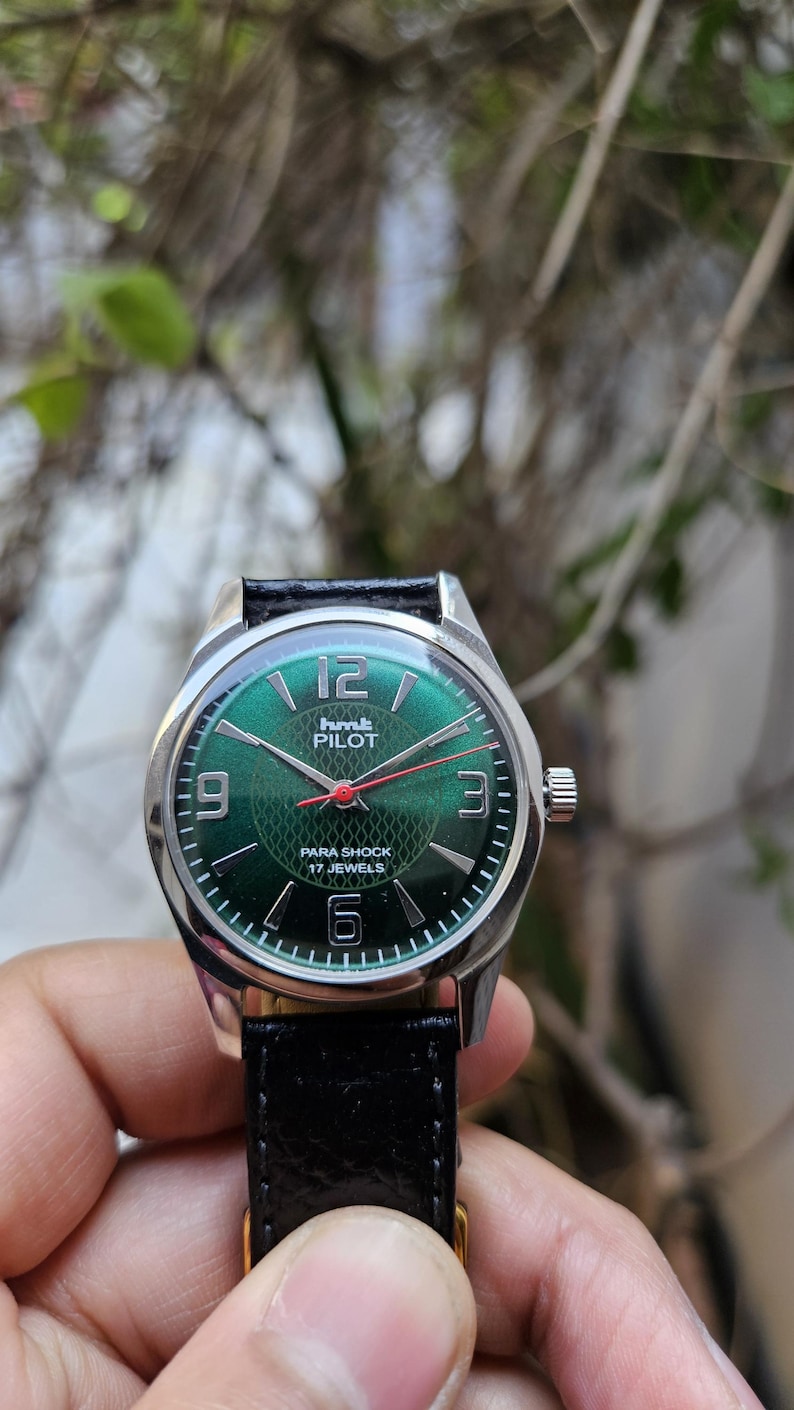 May include: A green-faced wristwatch with a silver case and black leather band. The watch face has white numerals and a red second hand. The watch face also has the text "hmt PILOT PARA SHOCK 17 JEWELS".