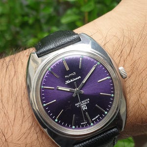 May include: A silver wristwatch with a black leather strap. The watch face is purple with silver hands and markers. The watch face also has the text "Kohinoor" and "Para Shock 17 Jewels" printed on it.