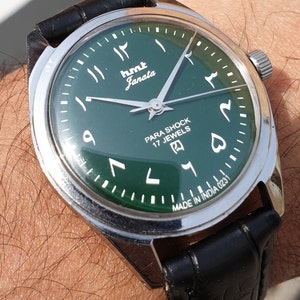 May include: A green-faced wristwatch with a black leather strap. The watch face has Arabic numerals and the text "HMT Janata" and "Para Shock 17 Jewels". The watch is made in India.