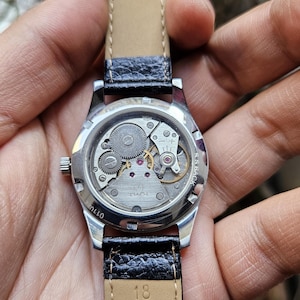 May include: A silver wristwatch with a black leather band. The watch face is visible and shows the inner workings of the watch. The band is labeled "Genuine Leather Co."