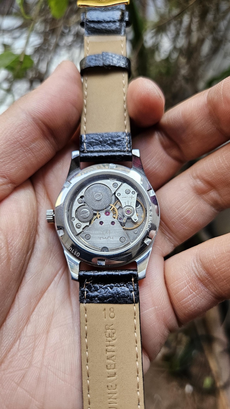 May include: A silver wristwatch with a black leather band. The watch face is visible and shows the inner workings of the watch. The band is labeled "Genuine Leather Co."
