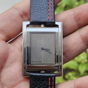 May include: A rectangular silver wristwatch with a gray face and black leather band. The band has red stitching. The watch face has the brand name "TITAN" printed on it. The watch is a classic accessory.