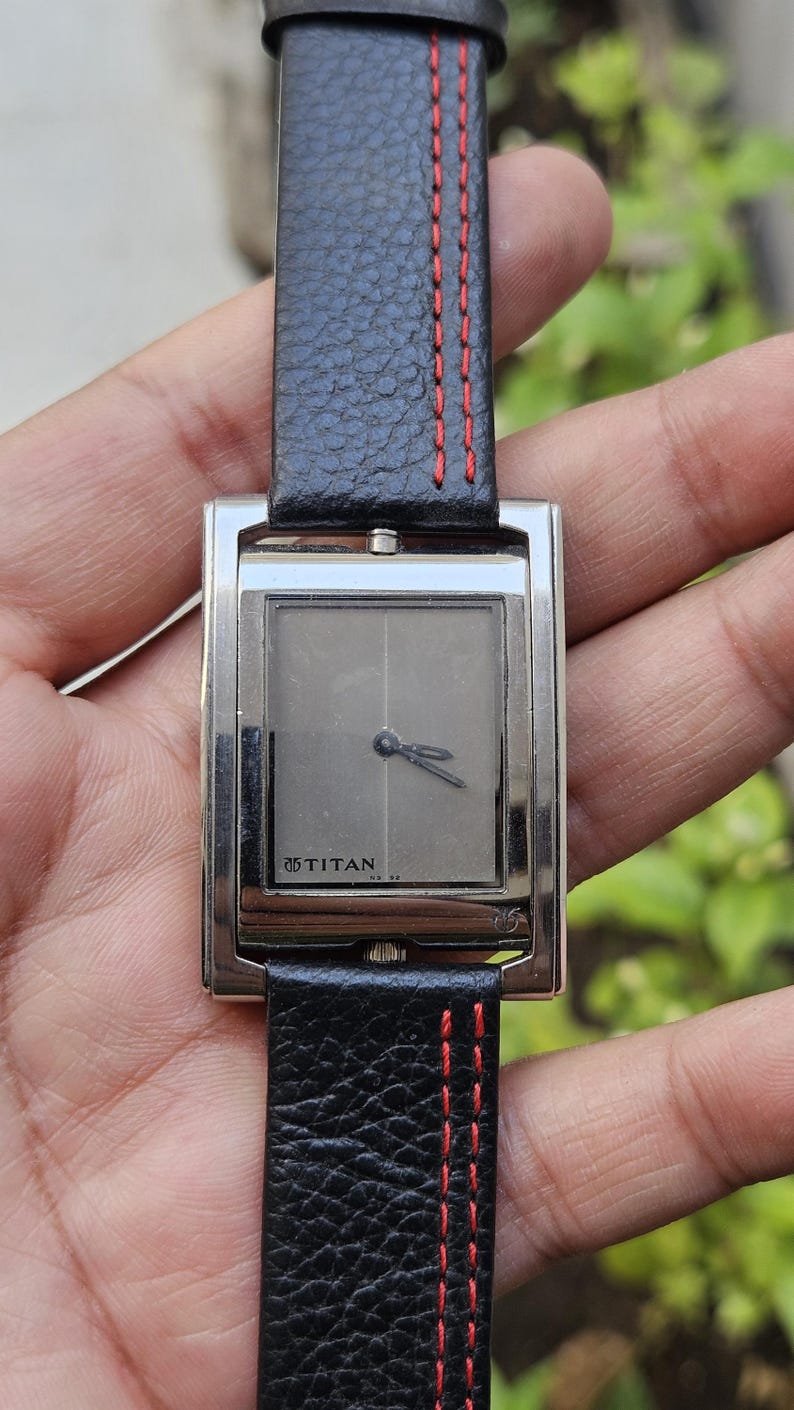 May include: A rectangular silver wristwatch with a gray face and black leather band. The band has red stitching. The watch face has the brand name "TITAN" printed on it. The watch is a classic accessory.