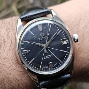 May include: A vintage Titus watch with a black dial, silver hands, and a date window at the 3 o'clock position. The watch has a stainless steel case and a black leather strap. The dial reads "25 Jewels Automatic" and "Swiss Made".
