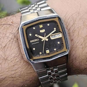Może przedstawiać: Zegarek Seiko Automatic w stylu vintage z czarną kwadratową tarczą i złotymi akcentami. Zegarek posiada wyświetlacz daty i dnia oraz bransoletę ze stali nierdzewnej. Na tarczy widnieje logo Seiko i napis "Automatic".