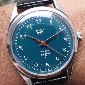 May include: A silver wristwatch with a teal face and black leather band. The watch face has white Arabic numerals and a red second hand. The watch face also has the text "humt janata" and "पैरा शाक १७ ज्वेल्स  四". The watch face also has the text "भारत में निर्मित ०२३१".