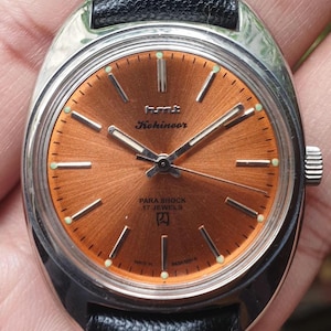 May include: A vintage wristwatch with a brown face and silver casing. The watch has a black leather strap and says "HMT Kohinoor Para Shock 17 Jewels" on the face.