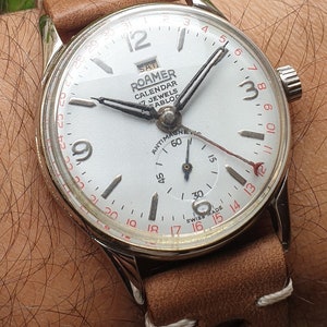 Może przedstawiać: Srebrny zegarek na rękę Roamer Calendar z białą tarczą i czarnymi wskazówkami. Zegarek ma brązowy skórzany pasek z białymi szwami. Tarcza zegarka zawiera tekst "Roamer Calendar 17 Jewels Sabloo" i "Antimagnetic 60".