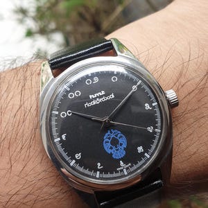 May include: A wristwatch with a black face and a silver-colored case. The watch face features white numerals and a blue emblem. The watch has a black leather band.