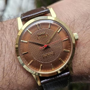 Könnte beinhalten: Eine goldfarbene Armbanduhr mit einem braunen Lederarmband. Das Zifferblatt der Uhr ist braun mit einem gemusterten Design und verfügt über goldfarbene Zeiger und Stundenmarkierungen. Das Zifferblatt der Uhr enthält auch den Text "PARA SHOCK 17 JEWELS" und das Wort "SONA".