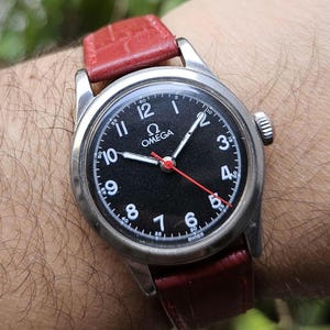 May include: A vintage Omega wristwatch with a black face, white numerals, and a red second hand. The watch has a stainless steel case and a red leather band. The watch is a classic timepiece.