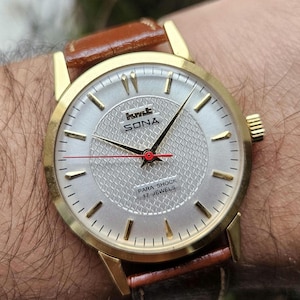 Könnte beinhalten: Eine goldfarbene Armbanduhr mit silbernem Zifferblatt und braunem Lederarmband. Das Zifferblatt zeigt den Markennamen "SONA" und den Text "PARA SHOCK 17 JEWELS". Die Zeiger der Uhr sind schwarz mit einem roten Sekundenzeiger.