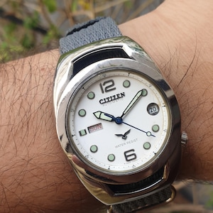 Peut inclure: Une montre-bracelet Citizen Automatic argentée avec un cadran blanc, des aiguilles noires et un bracelet en tissu gris. Le cadran de la montre comporte une fenêtre de date, une fenêtre de jour de la semaine et les mots "Water Resist" imprimés dessus.