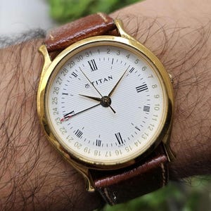 May include: A gold-tone analog wristwatch with a white textured dial, Roman numeral hour markers, and a date ring numbered 1 to 31. The watch features the Titan logo, gold hands, and a brown leather strap with visible stitching on the lugs.