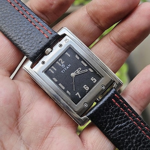 May include: A rectangular Titan wristwatch with a black face and silver-tone case. The watch features a black leather band with red stitching. The watch face displays the Titan logo and the numbers 1, 5, 6, 7, 11, and 12.