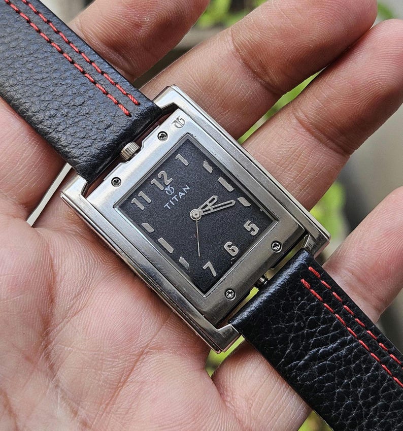 May include: A rectangular Titan wristwatch with a black face and silver-tone case. The watch features a black leather band with red stitching. The watch face displays the Titan logo and the numbers 1, 5, 6, 7, 11, and 12.