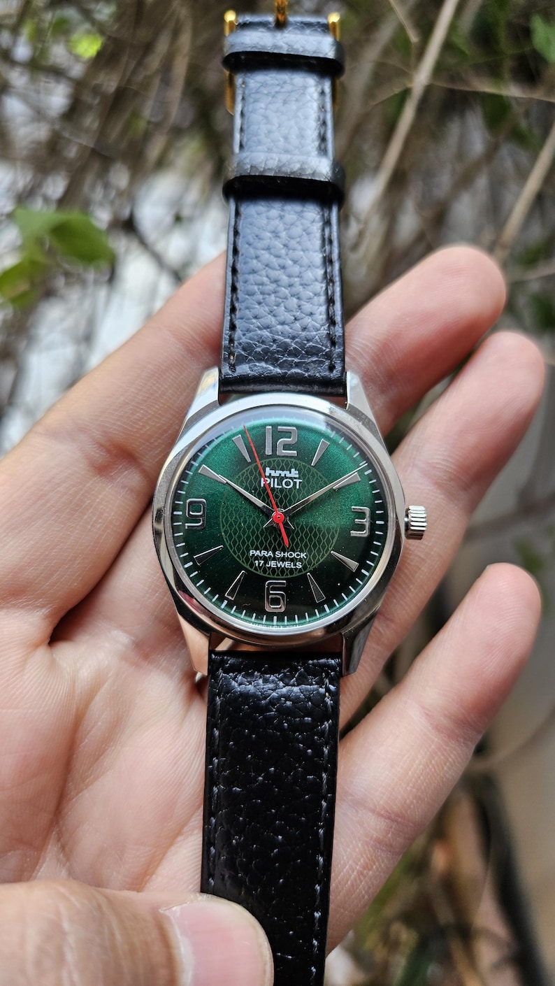 May include: A black leather wristwatch with a silver case and a green face. The face has white numbers and a red second hand. The watch says "Humt Pilot Para Shock 17 Jewels".