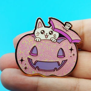 May include: A pink glitter pumpkin with a purple jack-o'-lantern face and a white cat peeking out of the top. The cat is wearing a pink hat with a gold trim.