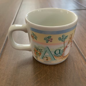 Peut inclure: Une tasse en céramique blanche avec un motif coloré représentant la lettre A, le mot "pommes" et un lapin. Le motif est dans un schéma de couleurs pastel avec du vert, du jaune, du bleu et du rose.