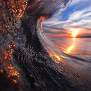 May include: A close-up view of a wave breaking, showing the sunset through the water. The water is a deep blue with orange and yellow hues from the sunset.