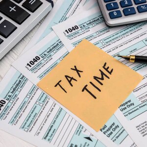 Può includere: Un post-it giallo con le parole "TAX TIME" scritte in nero su una pila di moduli fiscali americani, una calcolatrice e una penna.