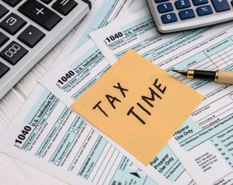 May include: A yellow sticky note with the words "TAX TIME" written in black marker on top of a stack of US tax forms, a calculator, and a pen.