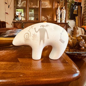 May include: A white, abstract sculpture with a rounded shape and a central opening. The sculpture features etched grey designs of figures and symbols. The piece is displayed on a wooden surface, showcasing its unique form and artistic detail.