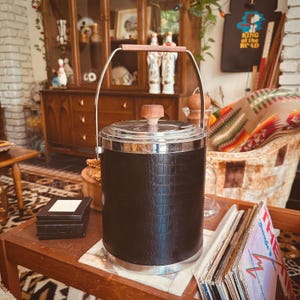 Puede incluir: Una cubitera vintage con exterior negro texturizado y borde plateado. Tiene un asa y un pomo de tapa de madera. La cubitera es cilíndrica y está sobre una mesa de madera, lo que sugiere un bar o un ambiente de entretenimiento.