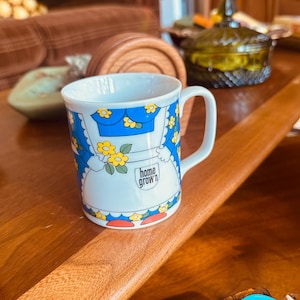 May include: White ceramic mug with a blue floral design and the words "Home Grown" printed on the front. The mug features a handle and is decorated with yellow flowers and a blue apron design.