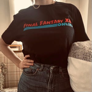 Puede incluir: Una camiseta negra de manga corta con el texto FINAL FANTASY XIV ONLINE impreso en rojo y azul en el pecho, con tres líneas horizontales azules debajo del texto. La camiseta se combina con unos vaqueros de mezclilla azul oscuro.