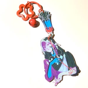 Puede incluir: Un llavero colorido con un personaje de estilo anime con una capa negra, detalles morados y azules y una cuerda roja. El llavero incluye una campana roja, un dije en forma de flor y un diseño de carta.