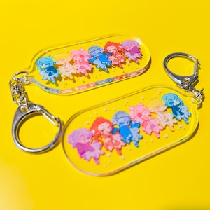 May include: Two clear acrylic keychains with cartoon character designs. Each keychain has a yellow background and a silver metal clasp. The characters are in various colors, including pink, blue, and purple.